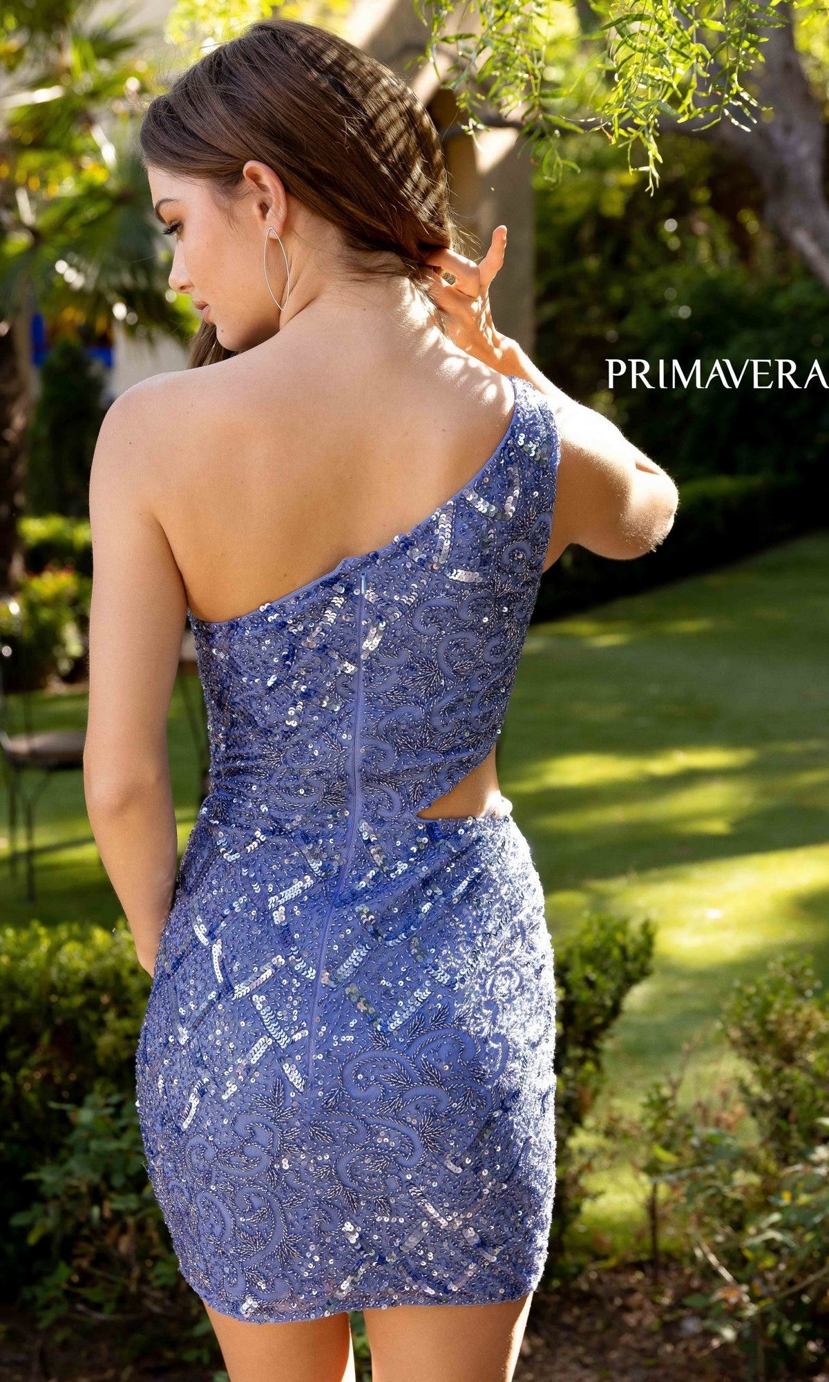 A woman in the Primavera 3504B One-Shoulder Short Party Dress stands outdoors in a garden, facing away from the camera and touching her hair.